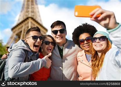 people, travel, tourism, friendship and technology concept - group of happy teenage friends taking selfie with smartphone and showing thumbs up over paris eiffel tower background