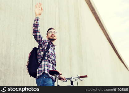 people, travel, tourism, gesture and lifestyle - happy young hipster man with fixed gear bike and backpack waving hand on city street