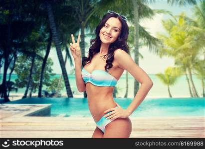 people, travel, tourism, gesture and summer concept - happy woman in bikini swimsuit showing victory hand sign over swimming pool and beach with palm trees background