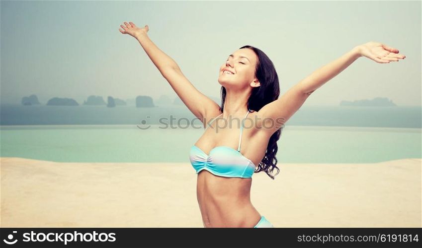 people, travel, tourism, happiness and summer concept - happy young woman in bikini swimsuit with raised hands looking up over infinity pool at sea side background