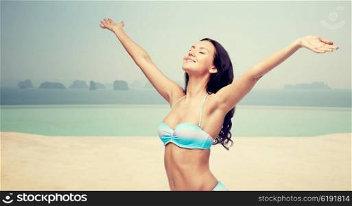 people, travel, tourism, happiness and summer concept - happy young woman in bikini swimsuit with raised hands looking up over infinity pool at sea side background