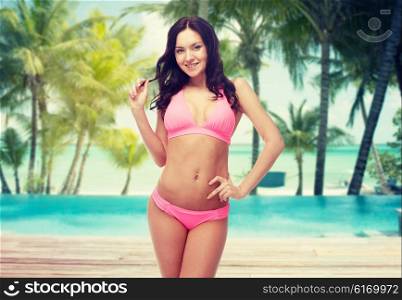 people, travel, tourism, summer and sexual concept - happy young woman posing in pink bikini swimsuit over swimming pool and beach with palm trees background