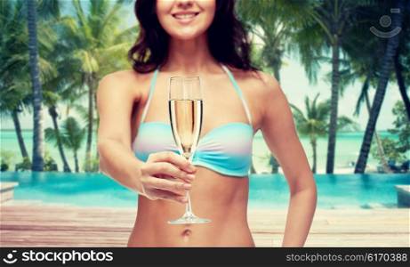 people, travel, tourism, summer holiday and celebration concept - happy young woman in bikini swimsuit drinking champagne at party over swimming pool and beach with palm trees background
