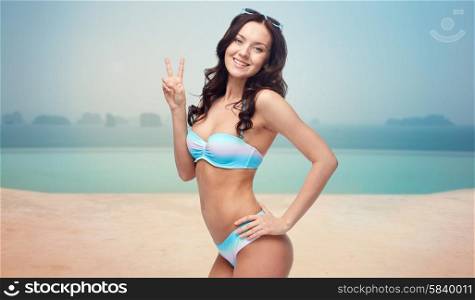 people, travel, tourism, summer holidays and celebration concept - happy woman in bikini swimsuit showing victory hand sign over infinity pool at sea side background