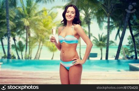 people, travel, tourism, summer holidays and celebration concept - happy young woman in bikini swimsuit drinking champagne at party over swimming pool and beach with palm trees background