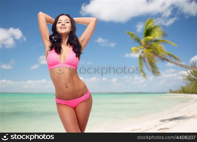 people, travel, tourism, swimwear and summer holidays concept - happy young woman posing in pink bikini swimsuit with raised hands over exotic tropical beach with palm trees background