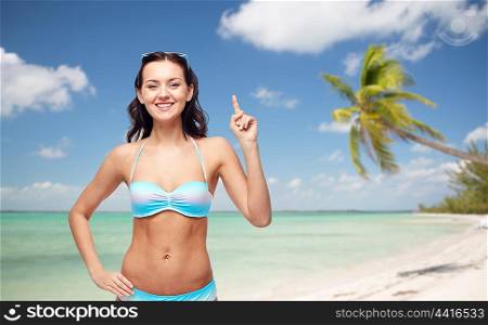 people, travel, tourism, swimwear and summer holidays concept - happy young woman in bikini swimsuit pointing finger up to something imaginary over exotic tropical beach with palm trees background