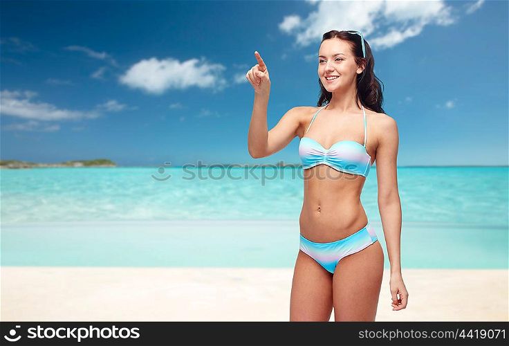 people, travel, tourism, swimwear and summer holidays concept - happy young woman in bikini swimsuit pointing finger to something imaginary over exotic tropical beach background