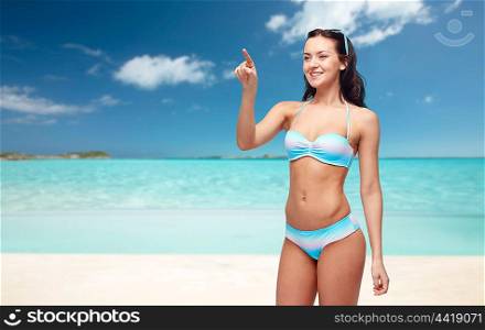 people, travel, tourism, swimwear and summer holidays concept - happy young woman in bikini swimsuit pointing finger to something imaginary over exotic tropical beach background