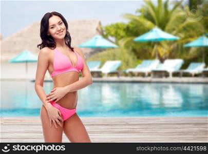 people, travel, tourism, swimwear and summer holidays concept - happy young woman posing in pink bikini swimsuit over swimming pool and sunbeds at exotic hotel resort background
