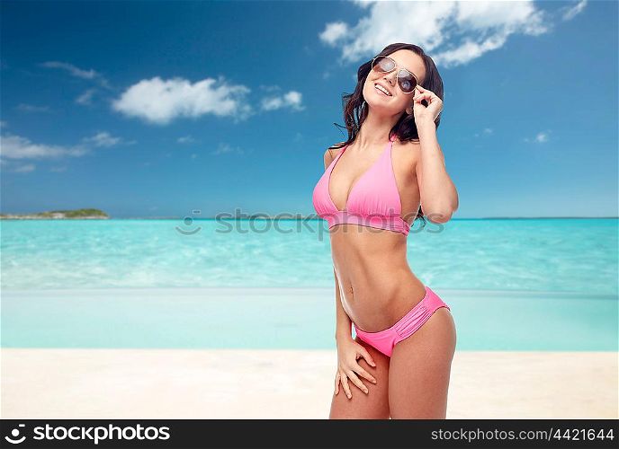 people, travel, tourism, swimwear and summer holidays concept - happy young woman in sunglasses and pink swimsuit over exotic tropical beach background