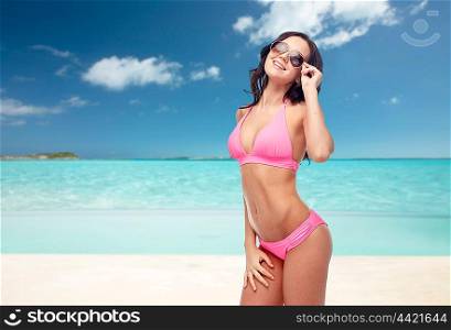 people, travel, tourism, swimwear and summer holidays concept - happy young woman in sunglasses and pink swimsuit over exotic tropical beach background