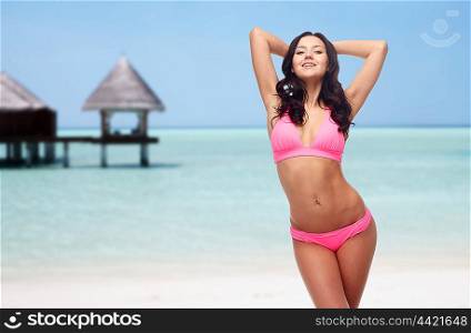 people, travel, tourism, swimwear and summer holidays concept - happy young woman posing in pink bikini swimsuit with raised hands over exotic tropical beach with bungalow in sea background