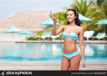 people, travel, tourism, swimwear and summer holidays concept - happy young woman in bikini swimsuit pointing to something imaginary over swimming pool and sunbeds at exotic hotel resort background