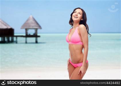 people, travel, tourism, swimwear and summer holidays concept - happy young woman in pink bikini swimsuit over exotic tropical beach with bungalow in sea background