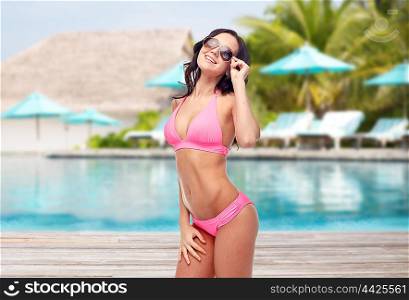 people, travel, tourism, swimwear and summer holidays concept - happy young woman in sunglasses and pink swimsuit over swimming pool and sunbeds at exotic hotel resort background