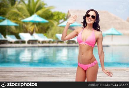 people, travel, tourism, swimwear and summer holidays concept - happy young woman in sunglasses and pink swimsuit waving hand over swimming pool and sunbeds at exotic hotel resort background