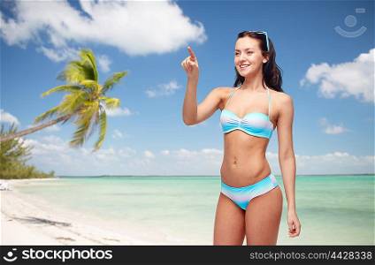 people, travel, tourism, swimwear and summer holidays concept - happy young woman in bikini swimsuit pointing finger to something imaginary over exotic tropical beach with palm trees background