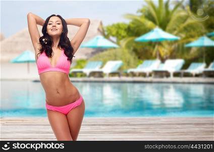 people, travel, tourism, swimwear and summer holidays concept - happy young woman posing in pink bikini swimsuit with raised hands over swimming pool and sunbeds at exotic hotel resort background