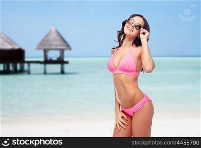 people, travel, tourism, swimwear and summer holidays concept - happy young woman in sunglasses and pink swimsuit over exotic tropical beach with bungalow in sea background