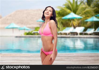 people, travel, tourism, swimwear and summer holidays concept - happy young woman in pink bikini swimsuit over swimming pool and sunbeds at exotic hotel resort background