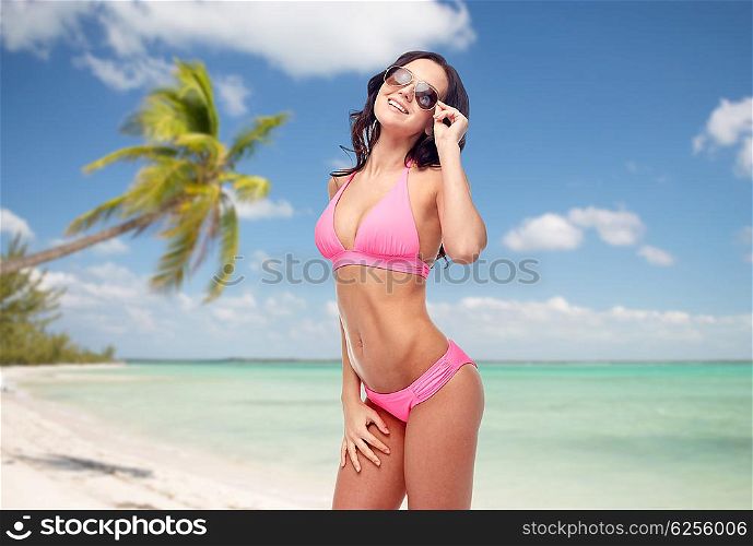 people, travel, tourism, swimwear and summer holidays concept - happy young woman in sunglasses and pink swimsuit over exotic tropical beach with palm trees background