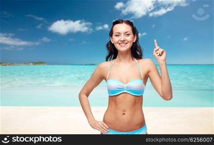 people, travel, tourism, swimwear and summer holidays concept - happy young woman in bikini swimsuit pointing finger up to something imaginary over exotic tropical beach background
