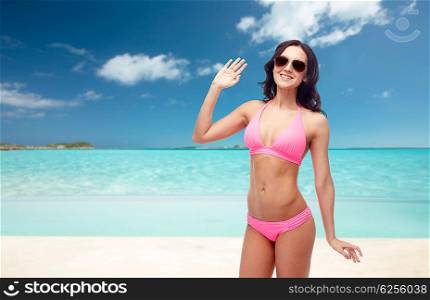 people, travel, tourism, swimwear and summer holidays concept - happy young woman in sunglasses and pink swimsuit waving hand over exotic tropical beach background