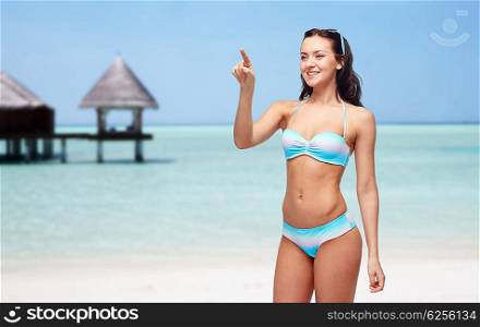 people, travel, tourism, swimwear and summer holidays concept - happy young woman in bikini swimsuit pointing finger to something imaginary over exotic tropical beach with bungalow in sea background