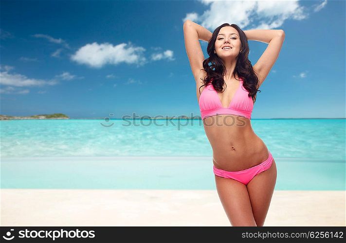 people, travel, tourism, swimwear and summer holidays concept - happy young woman posing in pink bikini swimsuit with raised hands over exotic tropical beach background