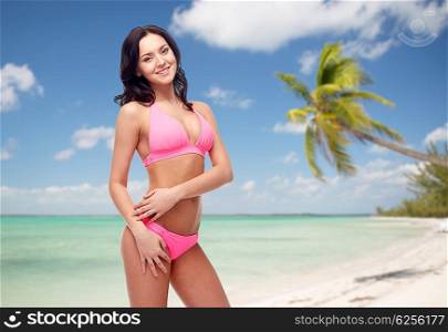 people, travel, tourism, swimwear and summer holidays concept - happy young woman posing in pink bikini swimsuit over exotic tropical beach with palm trees background
