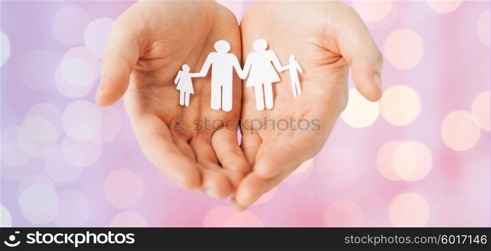 people, values and happiness concept - close up of man cupped hands showing paper family cutout over pink holidays lights background