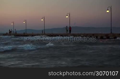 People walking along the pier with lanterns in evening. Rough sea with strong waves around