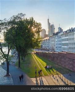 People walking and relaxing on green grass lawn at city embankment in evening sunlight, Frankfurt-am-Main, Germany
