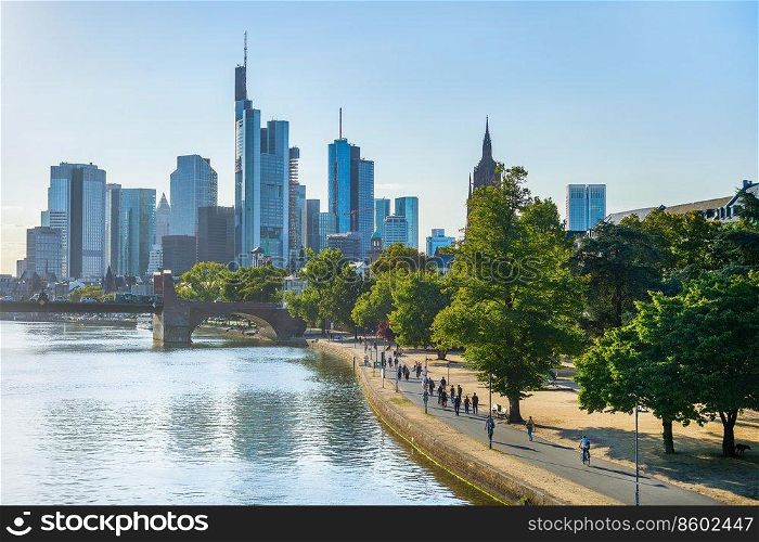 People walking by green Frankfurt embankment in evening sunlight, city skyline at background, Germany