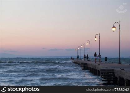 People walking on the pier in the evening. Lit lanterns standing along it. Rough sea with strong waves around