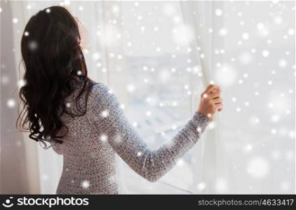 people, winter, christmas and home concept - close up of happy woman opening window curtains over snow