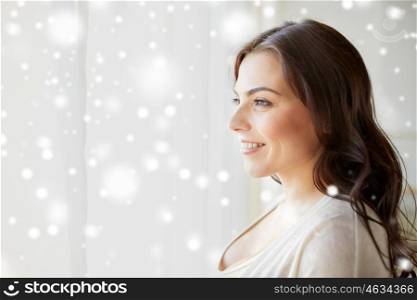 people, winter, christmas and leisure concept - happy young woman looking through window at home over snow