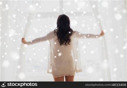 people, winter, christmas and morning concept - close up of happy woman opening window curtains at home over snow