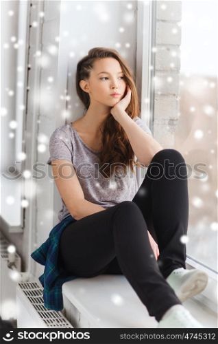 people, winter, christmas and teens concept - sad unhappy pretty teenage girl sitting on windowsill and looking through window over snow