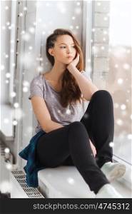 people, winter, christmas and teens concept - sad unhappy pretty teenage girl sitting on windowsill and looking through window over snow