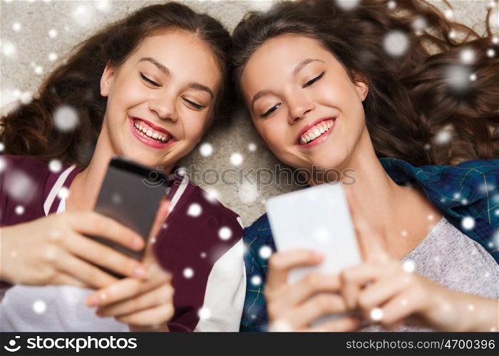 people, winter, christmas, technology and friendship concept - happy smiling pretty teenage girls or friends lying on floor with smartphones over snow