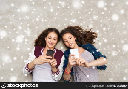 people, winter, christmas, technology and friendship concept - happy smiling pretty teenage girls or friends lying on floor with smartphones over snow