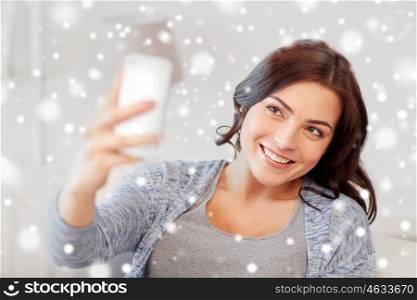 people, winter, christmas, technology and leisure concept - happy woman taking selfie with smartphone at home over snow