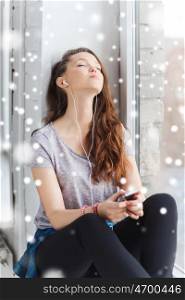 people, winter, christmas, technology and teens concept - sad pretty teenage girl sitting on windowsill with smartphone and earphones listening to music over snow