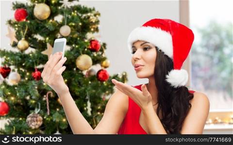 people, winter holidays and technology concept - beautiful woman in red santa hat taking selfie picture by smartphone and sending blow kiss to camera over christmas tree at home on background. woman in santa hat taking selfie by smartphone