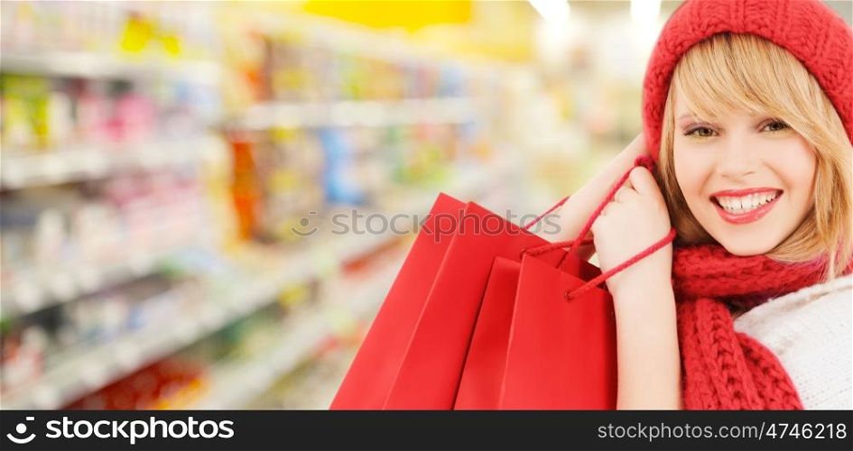 people, winter holidays, sale and consumerism concept - happy smiling woman in red hat and scarf with many shopping bags over supermarket background