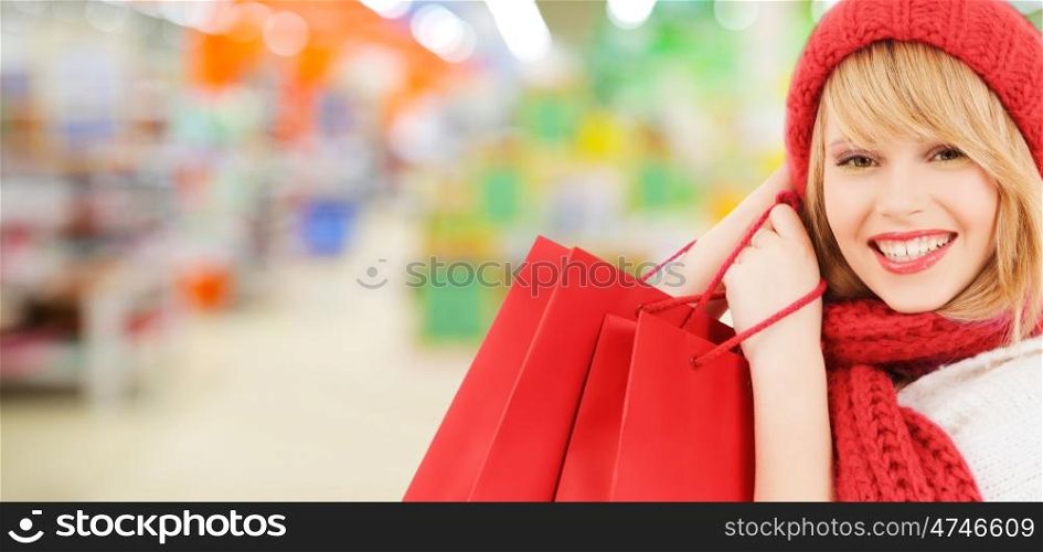 people, winter holidays, sale and consumerism concept - happy smiling woman in red hat and scarf with many shopping bags over supermarket background