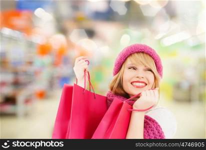 people, winter holidays, sale and consumerism concept - happy smiling woman in pink hat and scarf with many shopping bags over supermarket background