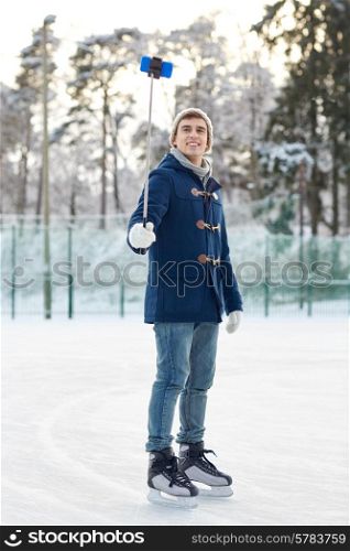 people, winter, technology and leisure concept - happy young man taking picture with smartphone selfie stick on ice skating rink outdoors